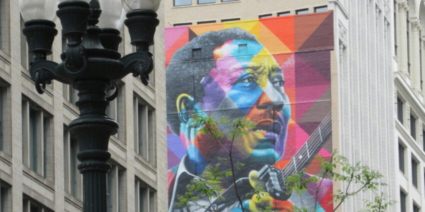 Colorful mural of a man playing a guitar, painted in a geometric, rainbow-toned style on the side of a city building, partially obscured by a tree and a black streetlamp.