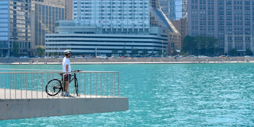 A cyclist with a helmet stands by his bike overlooking Lake Michigan with the Chicago skyline in the background.
