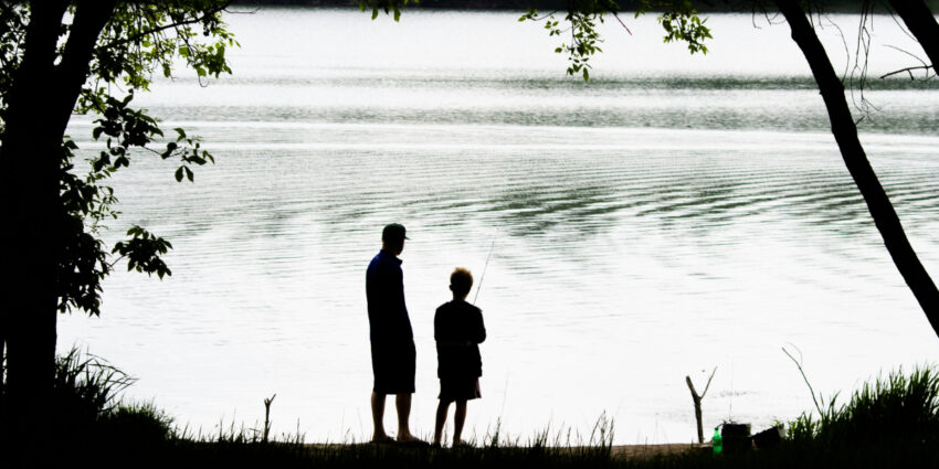 Silhouetted father and child fishing together on the shore of a calm lake, framed by trees and soft ripples on the water.